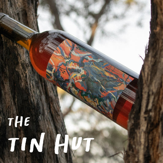 Wine bottle with colorful label leaning against a tree, 'The Tin Hut' branding visible